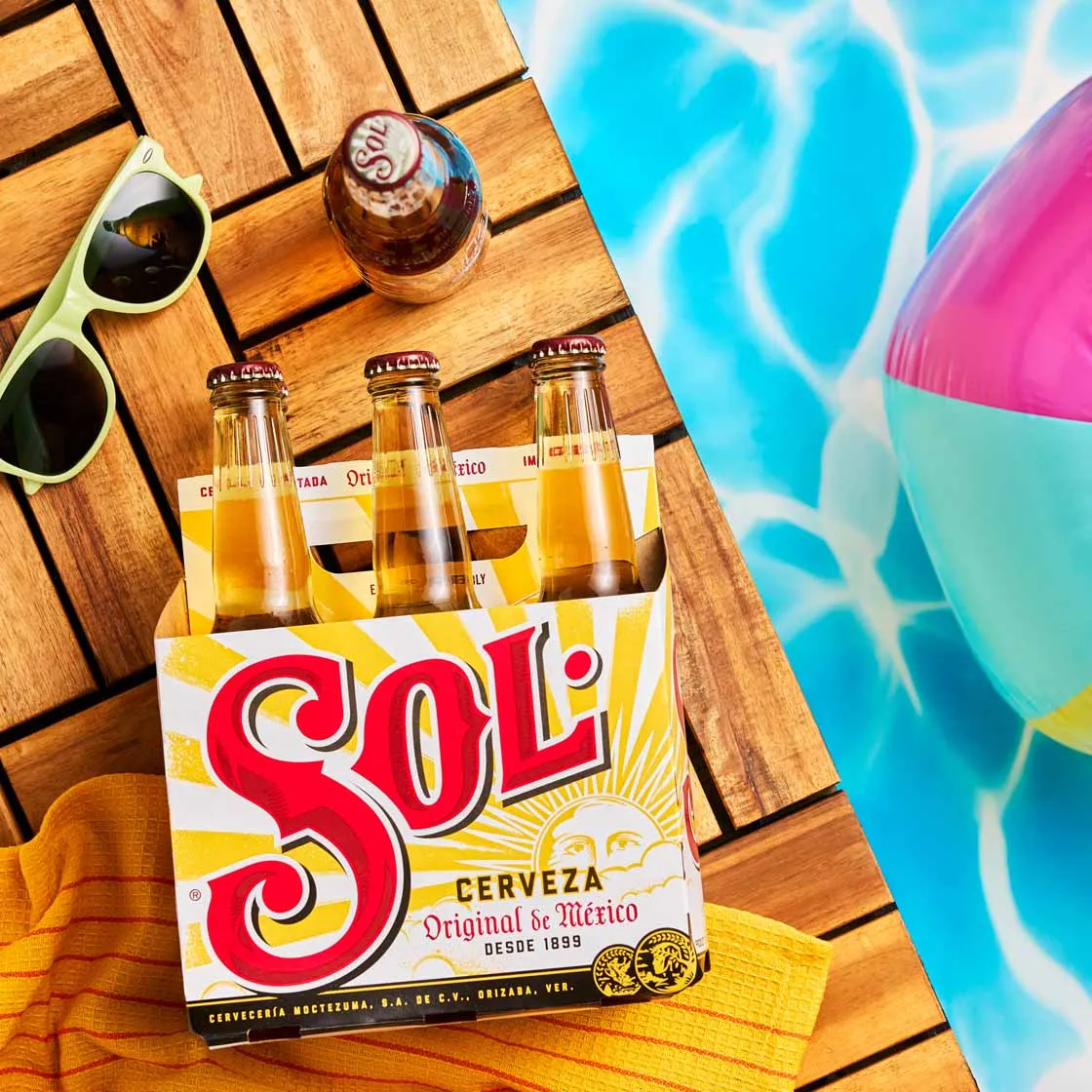 Sol beer six-pack on a poolside deck with sunglasses and a beach ball