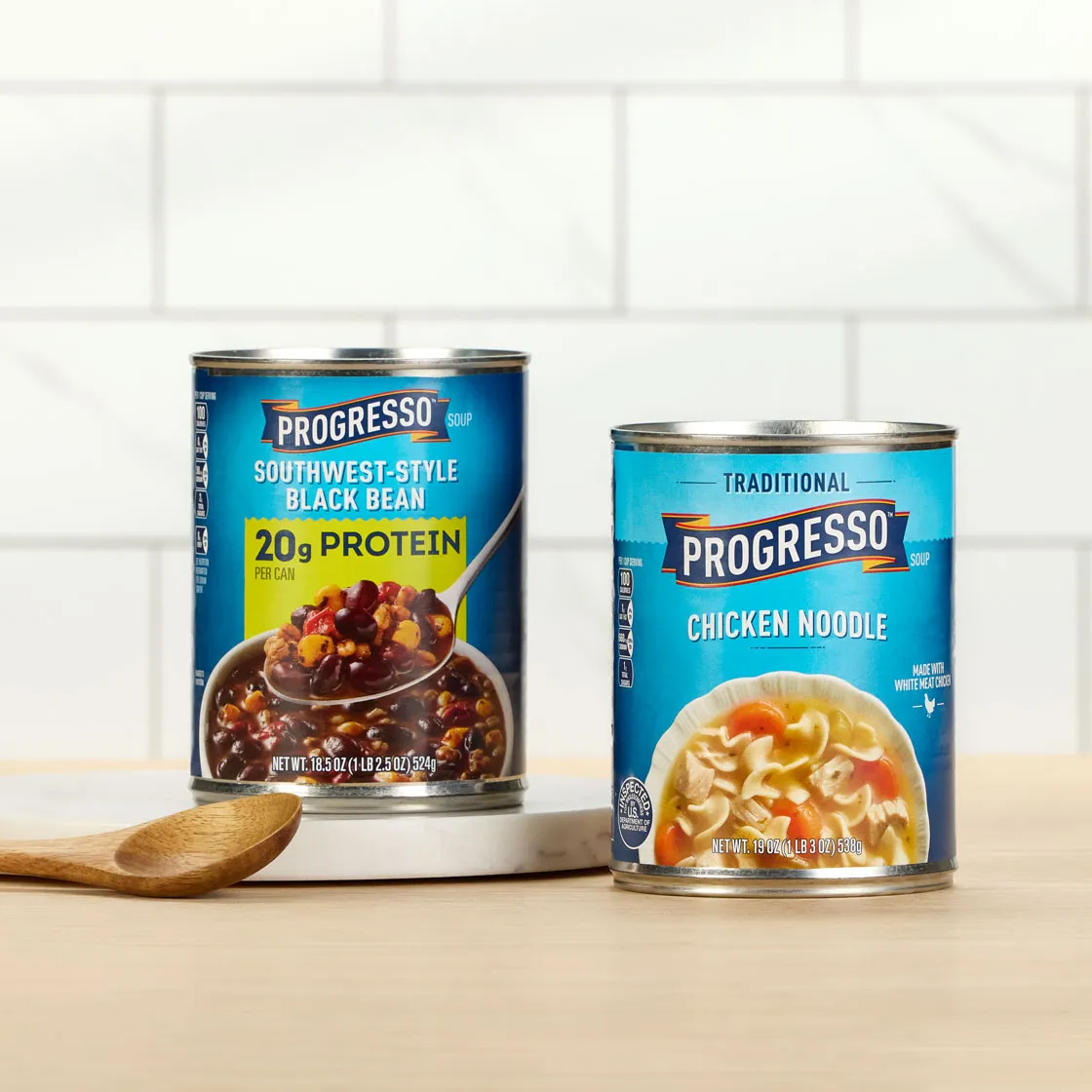 Two cans of Progresso soup on a kitchen counter with a wooden spoon