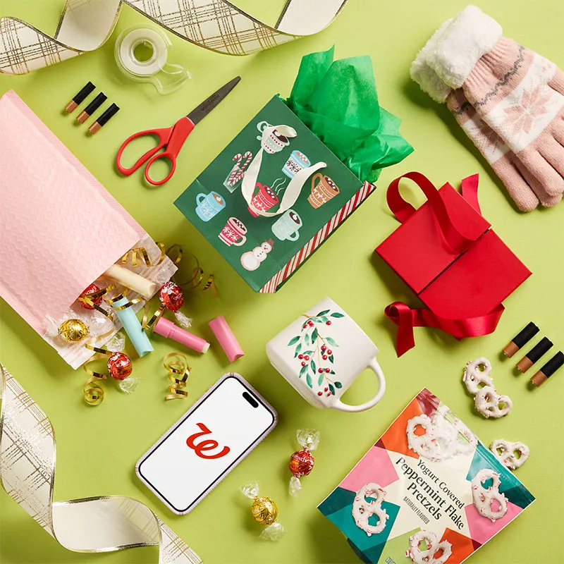 Holiday gifting flat lay with a phone, mugs, gloves, and wrapping on green