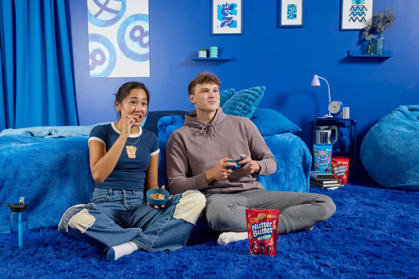 Two friends playing video games on a blue couch with snacks
